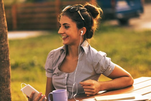 A snapshot captures a woman in a happy expression as she talks on the phone using a hands-free device. She is outdoors at a table in a park with greenery, organizing work or studying while sipping a h