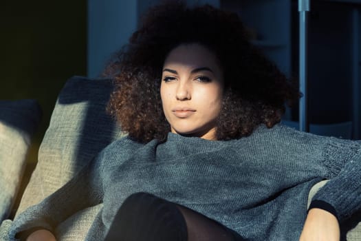 A fiercely confident young woman, successful and prosperous, lounges on her sofa in her spacious living room. Her curly hair, grey sweater, and beauty reflect her talent and dedication