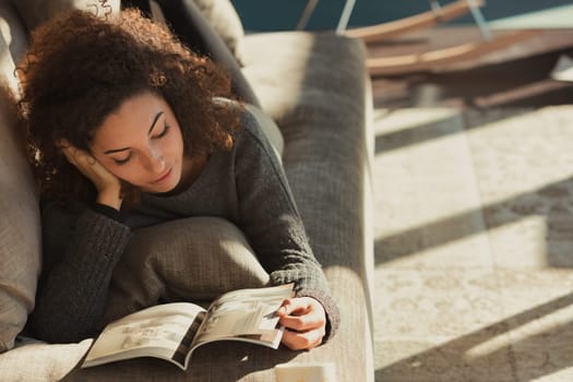 A relaxed young woman with curly hair lies belly-down on the couch in her spacious, sunlit living room, reading a magazine or catalog while enjoying her free time and having fun. Home sweet home