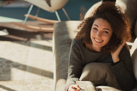 A relaxed young woman with curly hair is sprawled out on her stomach on the couch in her spacious and bright living room bathed in sunlight. She is reading a magazine or catalog with her head resting
