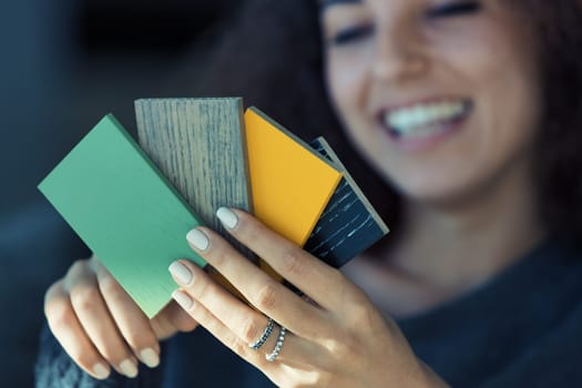 An ethnically diverse, beautiful woman with perfect teeth is seen on her couch, engrossed in selecting colors from a wood sample palette for her home. Her enjoyment in personalizing her lifestyle is e