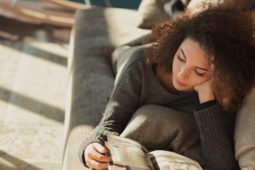 A relaxed young woman with curly hair is sprawled out on her stomach on the couch in her spacious and bright living room bathed in sunlight. She is reading a magazine or catalog with her head resting
