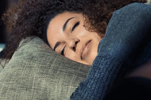 The sun warmly kisses a beautiful curly-haired girl's closed eyes as she cuddles on her gray couch, relishing in the warmth of her sweater and cozy home, dreaming or resting. She's stunning