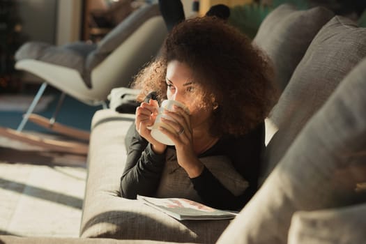 beautiful curly-haired woman is engrossed in reading a magazine. She takes a moment to thoughtfully sip a hot beverage, perhaps a herbal tea or regular tea, from her mug. She is comfortably settled on