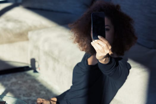 A smartphone in sharp focus, held by a young woman with curly hair. Taking selfies or exploring apps in her sun-drenched living room, her face is barely seen behind the screen