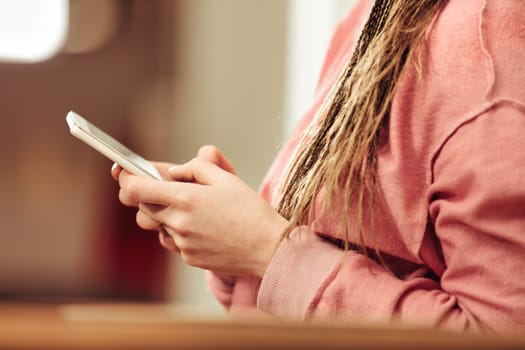 In warm light, close-up detail of female hands with smartphone at home with blurred background. She has an orange sweatshirt and box-braids