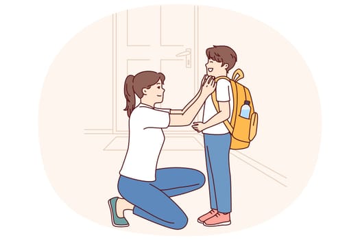 Single mother prepares young son of student in elementary school as trip to lessons and helps to put on briefcase kneeling down. Caring mother dresses teen boy for first day of school