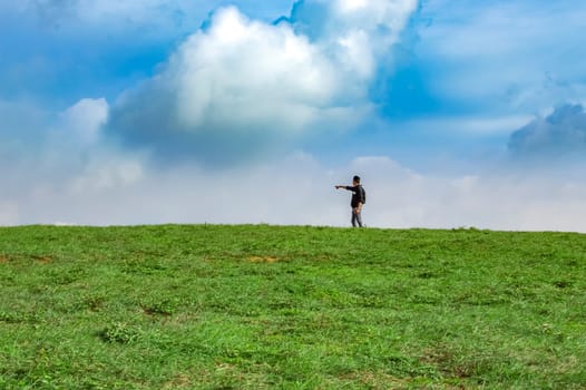 A backpacker on a hill with blue sky and copy space, man backpacking on a green hill with copy space, concept of successful man spreading his hands