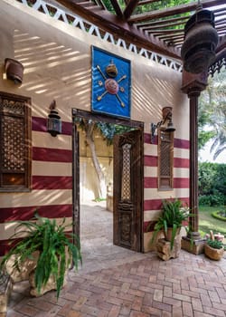 A beautifully crafted wooden door opens to a courtyard with red and white striped wall, wooden columns and lush greenery plant pots