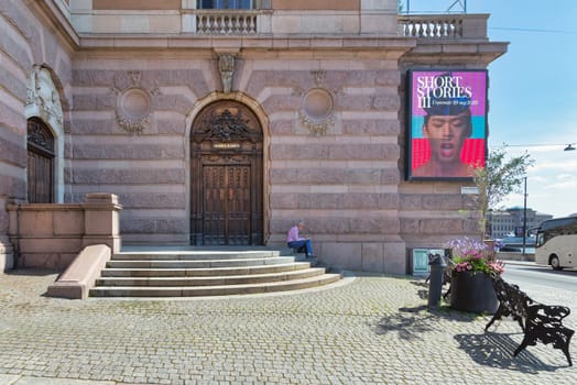 Stockholm, Sweden - June 26 2022: Elderly man reading a book at a staircase infront of brick stone wall with a billboard advertising Short Stories III and wooden door leading to the Royal Opera House