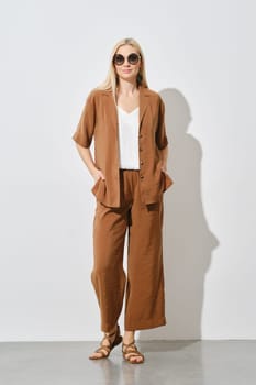 Cheerful blonde woman showcases a modern summer casual outfit. The loose brown suit and white inner top are paired with leather sandals