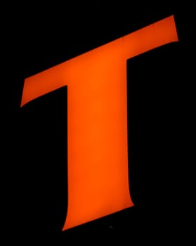 Captured from below is a striking, luminous 'T' set against a dark backdrop. Its audacious orange hue stands out vividly, contrasting sharply with its surroundings. Such a design choice might signify