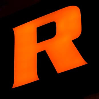 Captured from below is a striking, luminous 'R' set against a dark backdrop. Its audacious orange hue stands out vividly, contrasting sharply with its surroundings. Such a design choice might signify