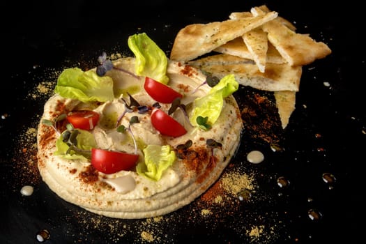 Indulge in a captivating hummus presentation, with smooth hummus forming a circular centerpiece adorned with juicy tomato slices, fresh green salad, and vibrant microgreens, all delicately dusted with paprika. Complemented by crispy pita bread slices, this platter promises a harmonious fusion of flavors and textures.