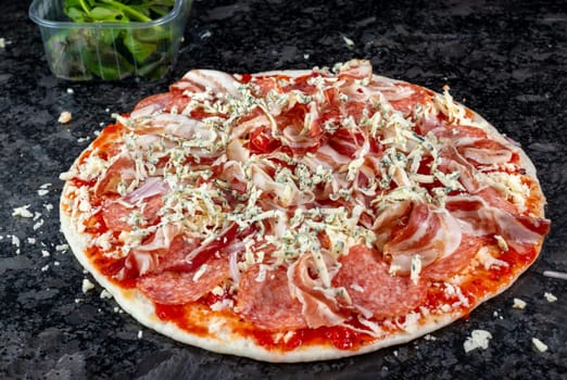 Behold a tantalizing pizza adorned with ham, salami, cheese, onions, and fresh herbs, resting on a granite kitchen countertop, eagerly awaiting its turn in the oven. Anticipation builds as the savory aromas fill the air