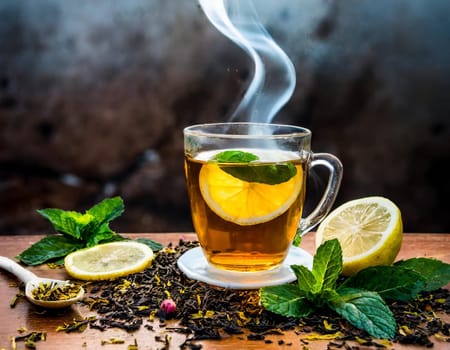 A clear glass cup of tea infused with lemon and mint leaves, steaming on a wooden table surr