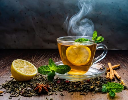 A clear glass cup of tea infused with lemon and mint leaves, steaming on a wooden table surr