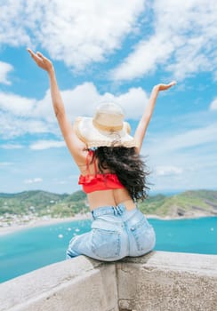 Lifestyle of woman on vacation sitting spreading hands to the beach. Tourist girl in hat spreading arms at a viewpoint looking at the beach