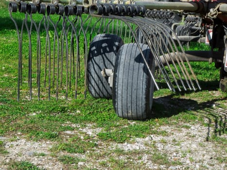 Partial view of two wheels and tines of a hay tedder on a gas surface in the sunshine.