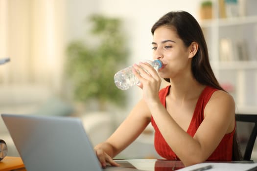 Student drinking water from bottle studying with a laptop at home