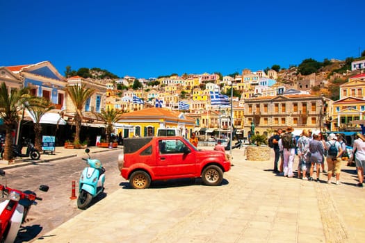 SYMI, Greece - June 03, 2021. Tuorists at the harbour of Symi Town. Colorful villas stretch up the steep slopes.