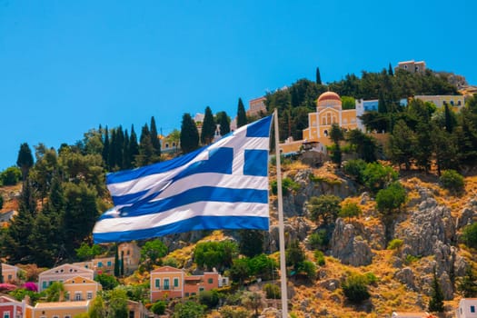 SYMI, Greece - JUN 03, 2021 A waving Greek flag in the harbour of Symi Town in front of colourful villas that rise up the steep mountain slopes.