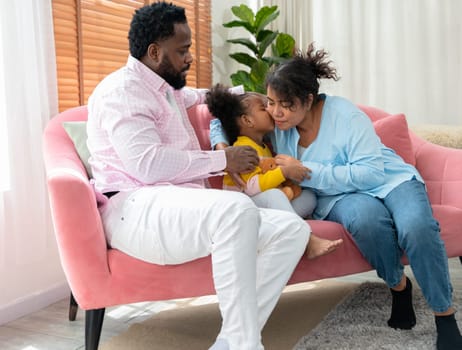 Little curly daughter kiss mother on cheek showing love and express caress while resting with parent on couch at home. Happy family, mother day, and sweet moment together concept