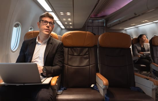 Young attractive businessman with eyeglasses working with laptop computer while sitting in airplane cabin. Internet connection on board service and business travel concept.