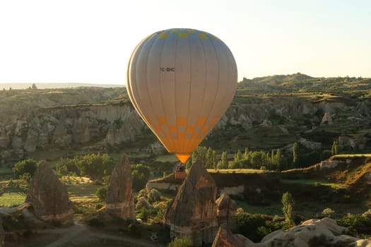 Yellow and white hot air balloon illuminated by the early morning sun while flying over the landscape of Cappadocia with its famous fairy chimneys, close to Göreme, Cappadocia, Turkey 2022