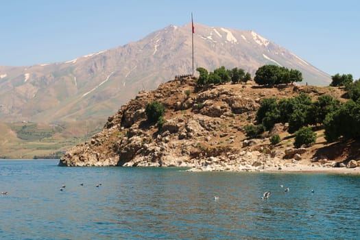 View from a boat onto Akdamar Island in the foreground with the red turkish flag on a pole and the snow covered Mount Artos in the background, Lake Van, Van Gölü, Turkey 2022