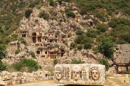 The ancient site of Myra with a stone relief with three theater masks in the front and the famous rock cut tombs in the background, near Demre, Turkey 2022
