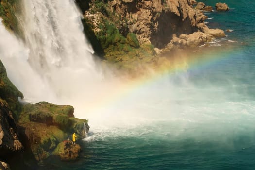 Fisherman in bright yellow raincoat trying to catch fish from a rock right next to the Düden Waterfalls in Antalya, Turkey 2022