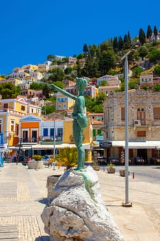 SYMI, Greece - JUN 03, 2021. Boy sculpture on Symi town promenade in the harbour in front of colorful villas that rise up the steep mountain slopes.