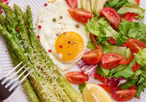 Round plate with cooked asparagus, fried egg, avocado and fresh vegetable salad on the table, top view