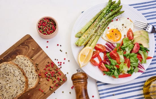 Round plate with cooked asparagus, fried egg, avocado and fresh vegetable salad on the table, top view