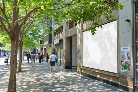 A blank digital billboard on a bustling city street, basked in sunlight and flanked by green trees.