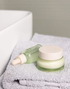 Translucent light green dropper bottle and closed cream jar on light grey folded bath towel near basin closeup, cosmetic mockup. Beauty and skincare routines with natural organic products