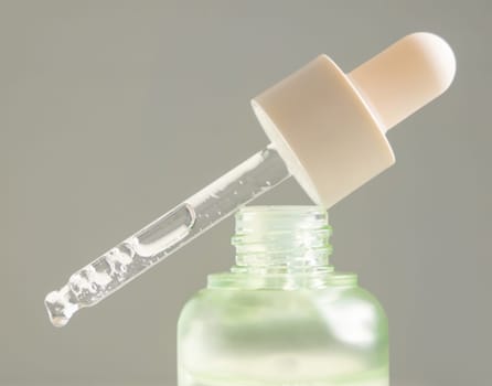 Drop falls from a pipette resting diagonally on a bottle filled with green liquid against gray backgound, close-up. Natural organic serum, gel, oil or other cosmetic product for everyday beauty routine
