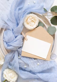 Card and envelope near light blue tulle fabric knot and cream roses on plates top view, copy space. Wedding stationery mockup. Romantic table place with flowers and pastel decor