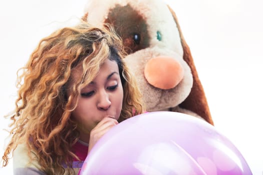 Young woman inflates a large pink balloon, with a stuffed dog toy looking over her shoulder, expressing a playful mood