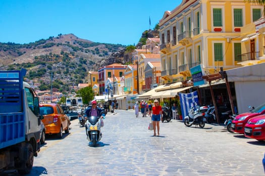 SYMI, Greece - June 03, 2021. Tourists at the harbour of Symi Town. Colorful villas stretch up the steep slopes.