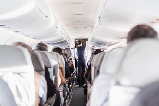 Interior of airplane with passengers on seats and stewardess in uniform walking the aisle, serving people. Commercial economy flight service concept