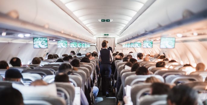 Interior of airplane with passengers on seats and stewardess in uniform walking the aisle, serving people. Commercial economy flight service concept