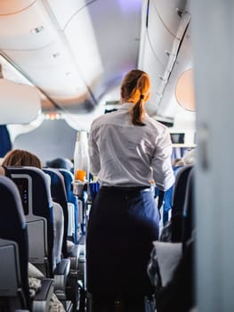 Interior of airplane with passengers on seats and stewardess in uniform walking the aisle, serving people. Commercial economy flight service concept