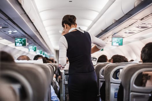 Interior of airplane with passengers on seats and stewardess in uniform walking the aisle, serving people. Commercial economy flight service concept