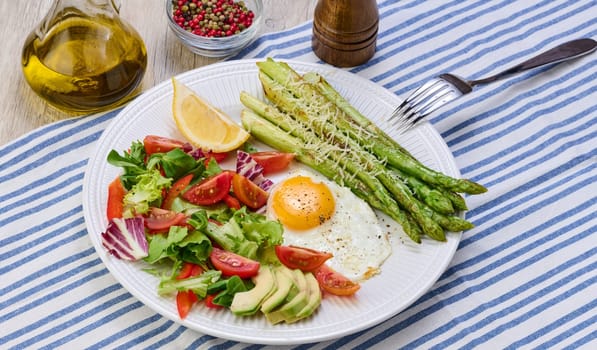 Round plate with cooked asparagus, fried egg, avocado and fresh vegetable salad on the table, top view