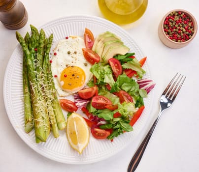 Round plate with cooked asparagus, fried egg, avocado and fresh vegetable salad on the table, top view