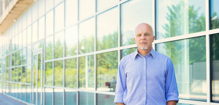 Mature man in light blue shirt poses outdoors, his demeanor calm and reflective against a contemporary glass facade