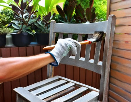 Garden chair with brush in protective gloves. Worker paints garden furniture. Renewal, renovation of wooden garden furniture