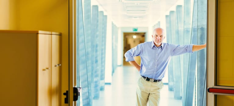 Standing in a bright hallway, an elderly man in a blue shirt leans against a door frame. He looks serious, thinking about future challenges like climate change, job losses, and power concentration